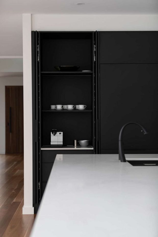 A modern minimalist kitchen viewed toward a tall matte-black built-in pantry and appliance cabinet with open shelving displaying cups and a coffee machine. A large pale island with an integrated dark faucet and sink occupies the foreground. White walls, warm timber flooring, and a partially visible timber door provide contrast.