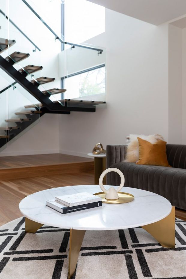 Bright contemporary living room with a sculptural floating staircase featuring dark structural stringers, timber treads and glass balustrades. The room has white walls, warm timber flooring, a dark channel-tufted sofa with mustard and cream cushions, and a round white marble coffee table with brass-toned legs. A large geometric black-and-white rug and double-height glazing reinforce the refined modern interior.