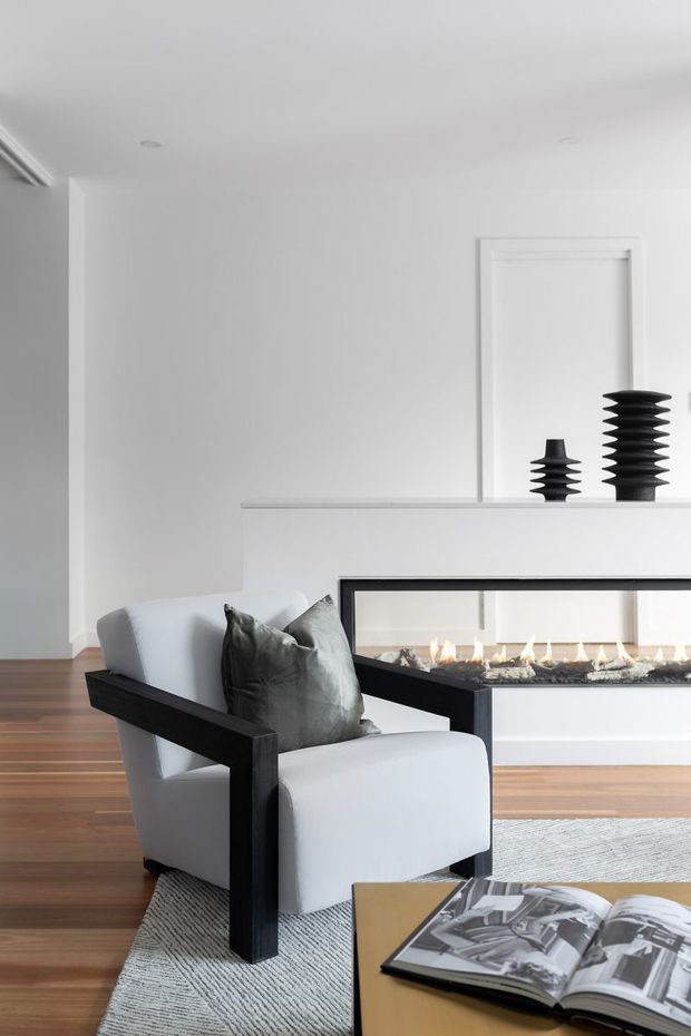 A refined contemporary living room with white walls, warm timber flooring, a long linear built-in fireplace with a black surround, and a sculptural white armchair with black timber arms in the foreground. A low coffee table with an open design magazine sits on a pale textured rug, while black decorative vases are displayed on the fireplace ledge.