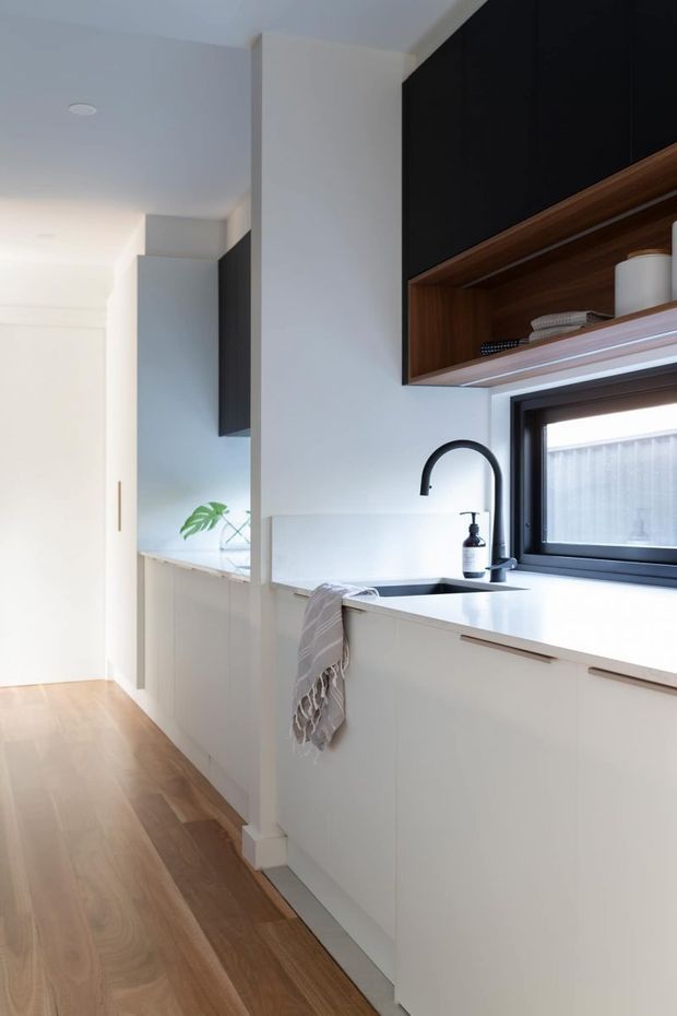 Bright, compact contemporary kitchen viewed along a hallway-like circulation space. The kitchen features white flat-panel cabinetry, a pale solid benchtop, integrated sink, matte black gooseneck tap, black-framed window, dark overhead cabinetry, and a warm timber open shelf. The room has white walls and ceiling with warm timber flooring, creating a clean, understated, high-contrast interior.