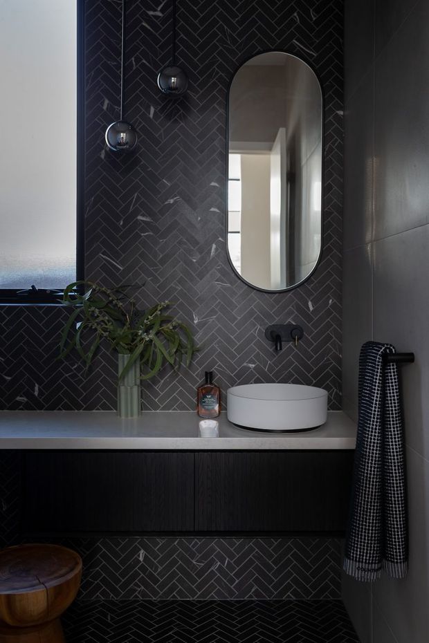 A moody, refined bathroom vanity with dark charcoal herringbone tile wrapping the wall and floor, a floating dark timber vanity with a pale stone benchtop, round vessel basin, black wall-mounted tapware, and a tall arched mirror. Globe wall lights, a draped patterned towel, and a small plant add detail. A partial timber toilet is visible at lower left.