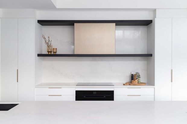 A bright, minimalist contemporary kitchen wall with white full-height cabinetry, a pale stone or engineered-stone benchtop and splashback, dark open shelving, and integrated black cooking appliances. Small timber and brass-toned decorative objects add subtle warmth.