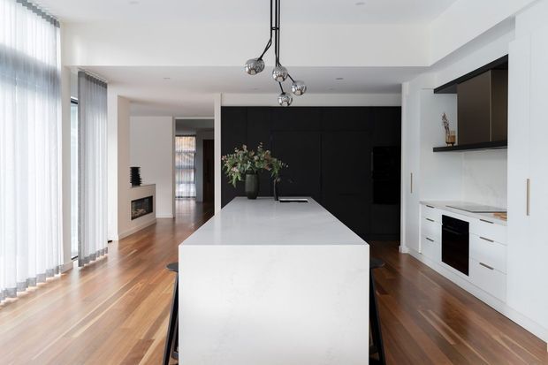 A bright contemporary open-plan kitchen with a large white island, white built-in cabinetry, black feature cabinetry and shelving, integrated appliances, and dark pendant lighting. Warm timber flooring extends through the adjoining living area, which includes a small fireplace and expansive sheer-covered glazing.