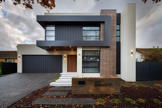 Street-facing exterior photograph of a contemporary two-storey residence with a strong geometric composition. The facade combines dark vertical cladding and garage door panels, smooth white rendered walls, extensive black-framed glazing, and a prominent vertical brick feature wall and planter. A covered entry sits beside the double garage, with paved paths, dark fencing, and minimal front landscaping.