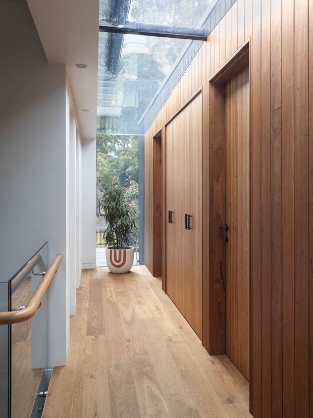 A bright, narrow residential hallway or landing with warm timber flooring and extensive vertical timber wall panelling and doors on the right. A glazed overhead roof section and full-height glazing at the end bring in abundant daylight and views of greenery. On the left is a staircase with a timber handrail and minimalist white walls. The space has a clean, contemporary design with strong natural-material detailing and built-in timber joinery.
