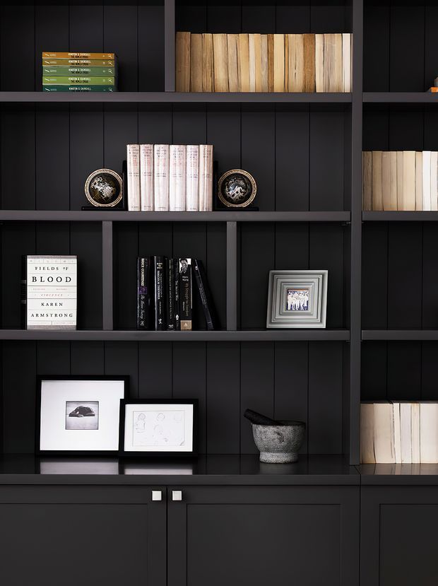 Close-up view of a dark, built-in shelving unit with vertical timber wall panelling, matte black shelves and cabinetry, and neatly arranged books, framed artwork, decorative plates, a vessel, and small accessories. The composition is highly ordered and monochromatic with warm wood and neutral accents.