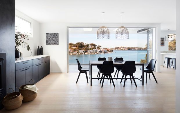Bright contemporary open-plan dining area with a large timber dining table and black chairs, a dark built-in sideboard, sculptural pendant lights, and expansive glazing overlooking a calm waterfront setting. The space has pale timber flooring, white walls, and a clean, minimalist coastal feel.