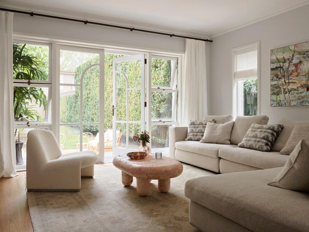 Bright, softly styled living room with large multi-pane windows and glazed French doors opening to a leafy garden. The room features an off-white sectional sofa, sculptural upholstered armchair, rounded wood coffee table, neutral patterned rug, white curtains, and abstract artwork in a calm, refined palette.