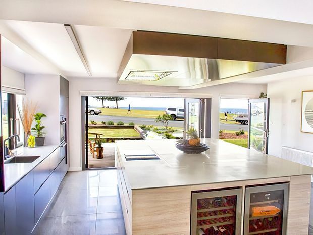 Bright contemporary kitchen with a large central island, integrated wine fridges, sleek cabinetry, and wide open glazing leading to a coastal outdoor view. The space features white surfaces, dark grey cabinetry, reflective stainless-steel detailing, and abundant natural light.