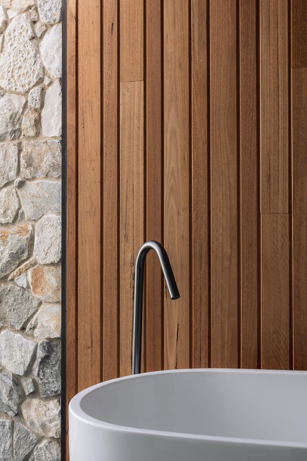 Close-up architectural detail of a freestanding white bathtub with a tall matte black floor-mounted faucet, set against vertical dark timber wall panelling and an adjacent rough natural stone wall. The composition highlights warm wood, textured masonry, and sculptural bathroom fixtures.