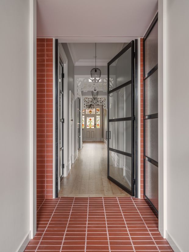 A narrow, formal residential entryway viewed from a tiled threshold, looking through an open black-framed glazed door into a long hallway. Red brick-toned tiled walls and floor surround the entrance, while the hall has pale timber flooring, white walls with decorative moulding, glass-panel doors, pendant lights, and a decorative stained-glass door at the far end. The composition emphasizes the corridor depth and layered doorways.