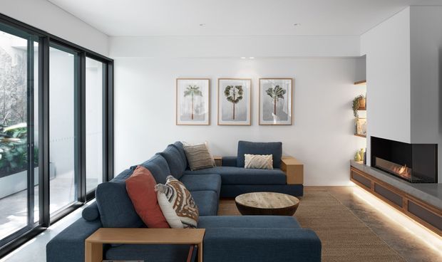 A bright contemporary living room with a large deep-blue sectional sofa arranged around a round coffee table. Full-height black-framed glazing lines the left side, while a built-in white media wall with open shelving, timber cabinetry, and a linear fireplace forms the right side. Three framed botanical artworks hang above the sofa, and warm concealed lighting accents the timber joinery.