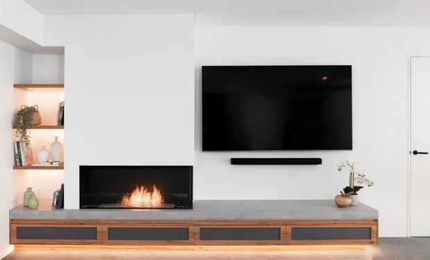A minimalist contemporary living room media wall featuring a large black television, linear fireplace with visible flames, and a long built-in timber entertainment unit with dark inset drawer fronts. Open white shelving with warm integrated lighting and decorative objects is visible to the left, while concealed LED lighting creates a soft glow beneath the joinery.