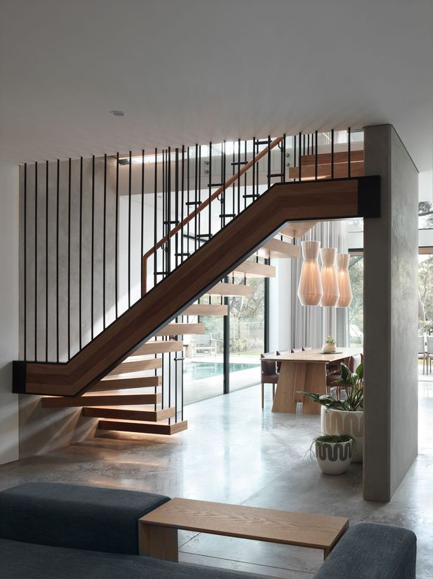 A spacious contemporary open-plan interior dominated by a sculptural timber staircase with open risers and slender black metal balustrade rods. The staircase spans across the room beside a substantial concrete-toned structural wall and column. A dining area with a timber table and pendant lights is visible beyond, along with floor-to-ceiling glazing, a polished concrete floor, indoor plants, and a glimpse of a swimming pool outside. The foreground includes a dark upholstered sectional sofa and timber coffee table.