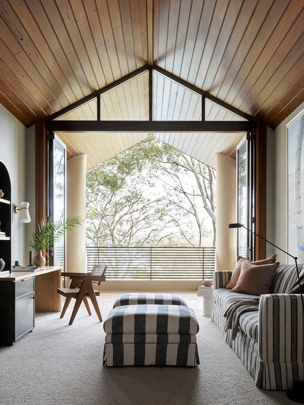 A warm, timber-lined living room with a dramatic pitched ceiling and exposed dark timber framing. The room opens through large glazed doors onto a covered balcony or terrace surrounded by trees. It features a striped upholstered sofa and ottoman, a small timber lounge chair, dark built-in shelving, a floor lamp, and pale carpet. The space feels relaxed, inviting, and connected to the outdoors.