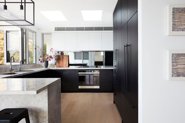 Bright contemporary kitchen with matte black full-height cabinetry, a large pale stone island, timber flooring, integrated appliances, and white walls. Skylights and extensive glazing provide abundant natural light, while a black linear pendant adds a geometric architectural feature.