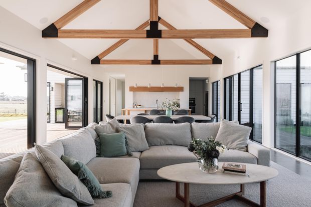 Bright open-plan living area with a large light-grey sectional sofa and round coffee table in the foreground, opening toward a kitchen and dining zone. Full-height black-framed glazing lines both sides, while exposed timber trusses with dark structural brackets form a vaulted ceiling. The palette is soft and neutral with muted green cushions and natural timber accents.