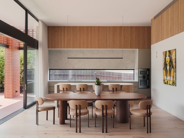 A refined contemporary dining area with a large solid-wood dining table and matching upholstered chairs, set beside full-height black-framed glazing. The room features warm timber wall panelling and cabinetry, a pale marble-look wall surface, integrated appliances, light timber flooring, and a linear suspended light. A brick courtyard is visible through the glazing, while a colourful artwork provides a small accent on the white wall.