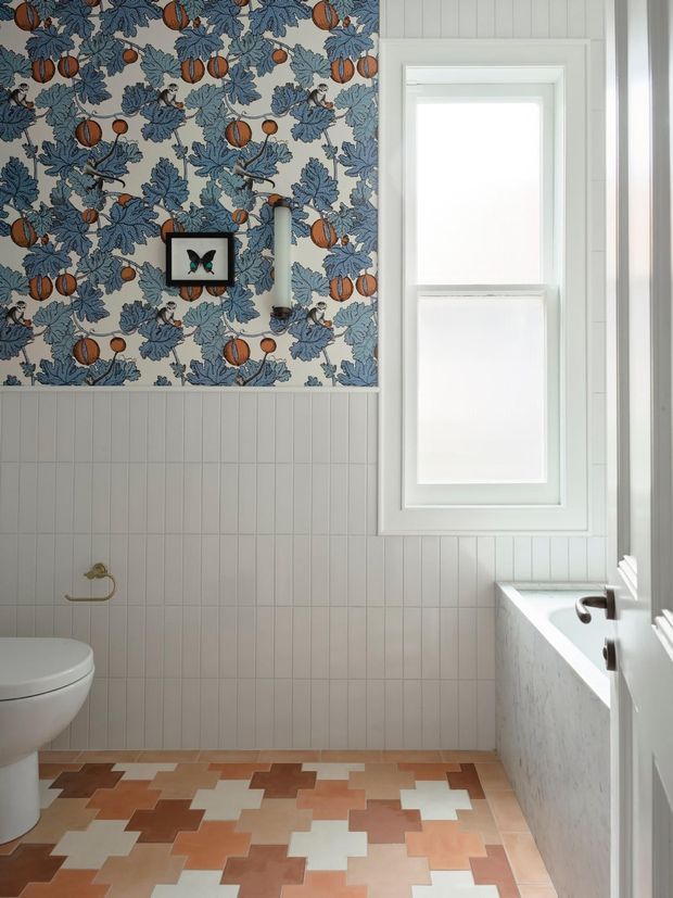 A bright bathroom with a white toilet, bathtub, framed recessed window, vertical white wall panelling, and a patterned upper wall featuring blue foliage and orange fruit motifs. The floor has a geometric patchwork of terracotta, brown, cream, and white tiles. A small framed butterfly artwork and brass toilet-roll holder add decorative detail.