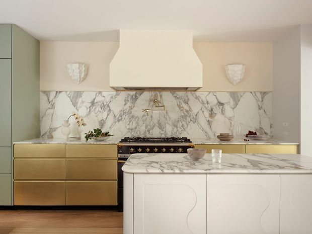 A refined, warm-toned kitchen featuring a large marble-look splashback and matching island benchtop, pale cream cabinetry, brushed brass drawer fronts and hardware, a statement range cooker, and a sculptural white rangehood. Decorative wall sconces and subtle styling create an elegant, luxurious interior.