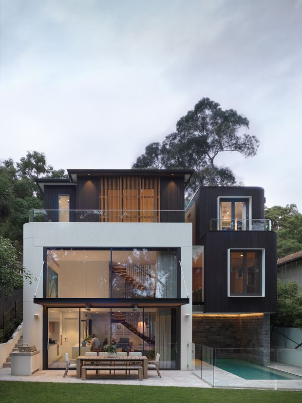 Photograph of a contemporary multi-level residential house set among mature trees. The facade combines dark vertical cladding, extensive floor-to-ceiling glazing, a white rendered rectangular volume, timber screening, glass balustrades, and stone masonry at the lower level. A covered outdoor dining area opens onto a lawn and a narrow swimming pool, with warm interior lighting visible through the windows.