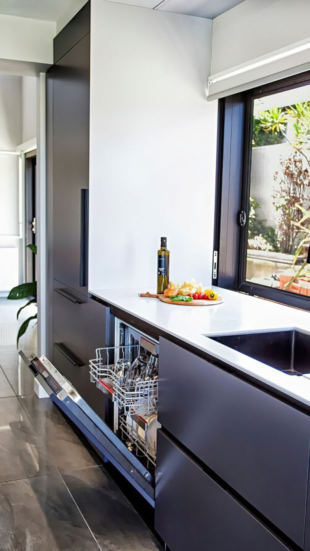 A contemporary galley kitchen with dark matte cabinetry, a bright white benchtop, integrated dishwasher drawers, a deep sink, and a large dark-framed window overlooking greenery. Polished dark floor tiles and abundant natural light create a clean, high-contrast interior.
