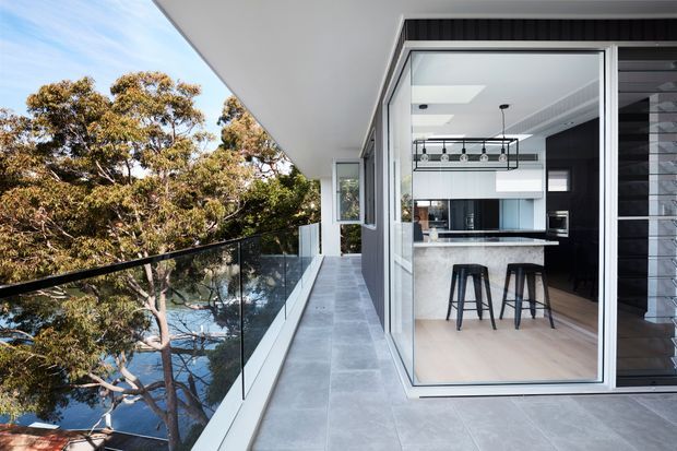 Contemporary apartment or house balcony opening directly into a modern kitchen. The image shows a long tiled balcony with frameless glass balustrading, extensive glazing, and a sleek white-and-black kitchen with an island, bar stools, and linear pendant lighting. Mature trees are visible beyond the balcony.