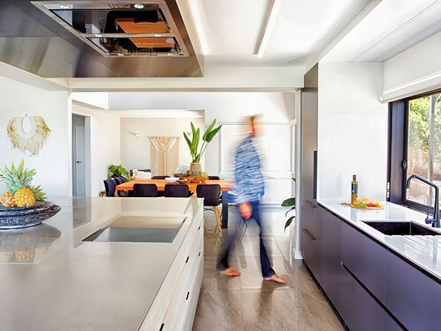 Bright contemporary open-plan kitchen viewed from the main work area, with a large pale stone island, integrated cooktop and ceiling-mounted rangehood, dark lower cabinetry, a long window over the sink, and a dining area beyond. The space has white walls and ceilings, polished light stone surfaces, timber-toned flooring, and a clean, airy fit-out. A blurred person is moving through the room.