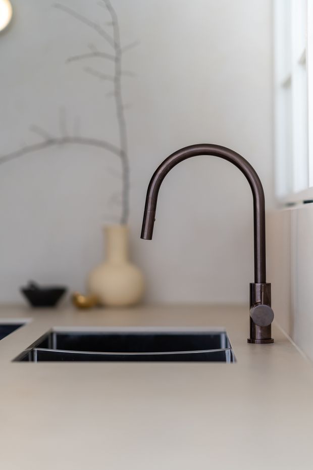 Close-up of a minimalist kitchen sink area featuring a dark brushed-metal gooseneck tap, integrated black sink, and pale smooth benchtop. A neutral wall, window, and softly blurred decorative vase form the background.