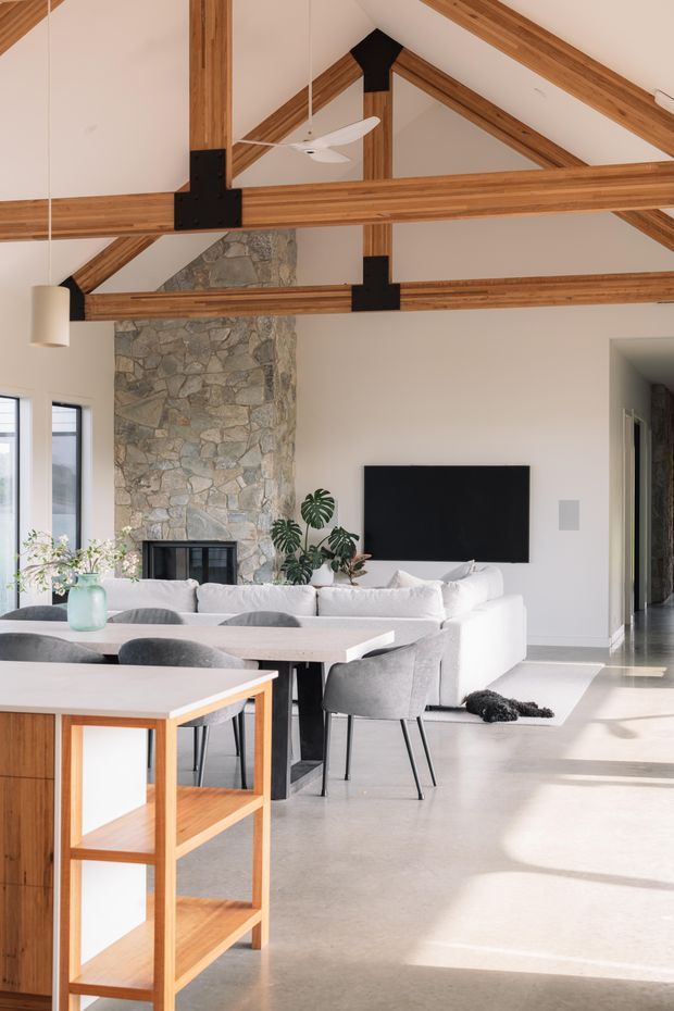 Bright open-plan living and dining space with a dramatic vaulted ceiling, exposed timber roof trusses, black metal connectors, and a tall natural stone fireplace wall. A white sectional sofa faces a wall-mounted television, while a light dining table with grey upholstered chairs and timber shelving occupies the foreground. Large windows bring in strong natural light, creating a warm contemporary-rustic atmosphere.