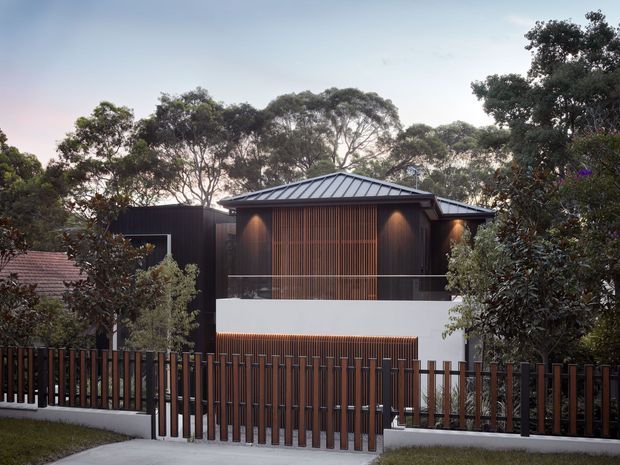 Exterior view of a contemporary two-storey residence set among mature trees. The house features dark vertical cladding, a low pitched metal roof, a prominent timber batten screen across the upper facade, white rendered walls, balcony glazing, and a matching vertical timber-and-metal front fence and gate.