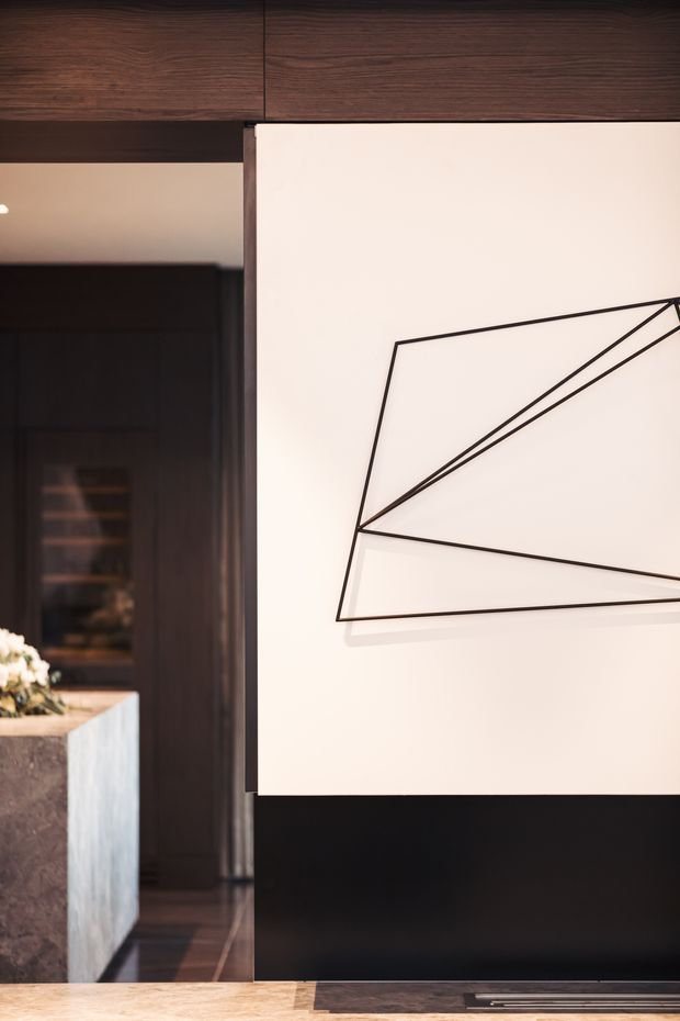 Close-up interior view of a refined dark-wood and black built-in wall with a large pale panel featuring minimalist geometric line artwork. A partial stone island or console and dark cabinetry are visible through the opening at left, creating a warm, contemporary architectural interior.