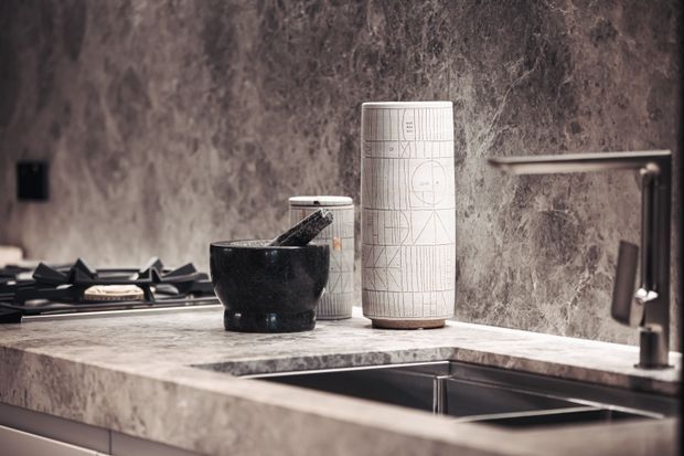 Close-up of a sophisticated kitchen countertop with a dark stone or marble-look backsplash and matching benchtop. A black bowl and patterned ceramic vessels sit beside a gas cooktop, while a deep stainless-steel sink and modern tap occupy the foreground.