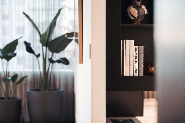 Close-up interior detail showing a dark built-in shelving unit with books and decorative objects beside tall indoor plants in planters, softened by sheer window curtains and natural light. The palette is predominantly black, white, grey, and muted green, creating a refined contemporary atmosphere.