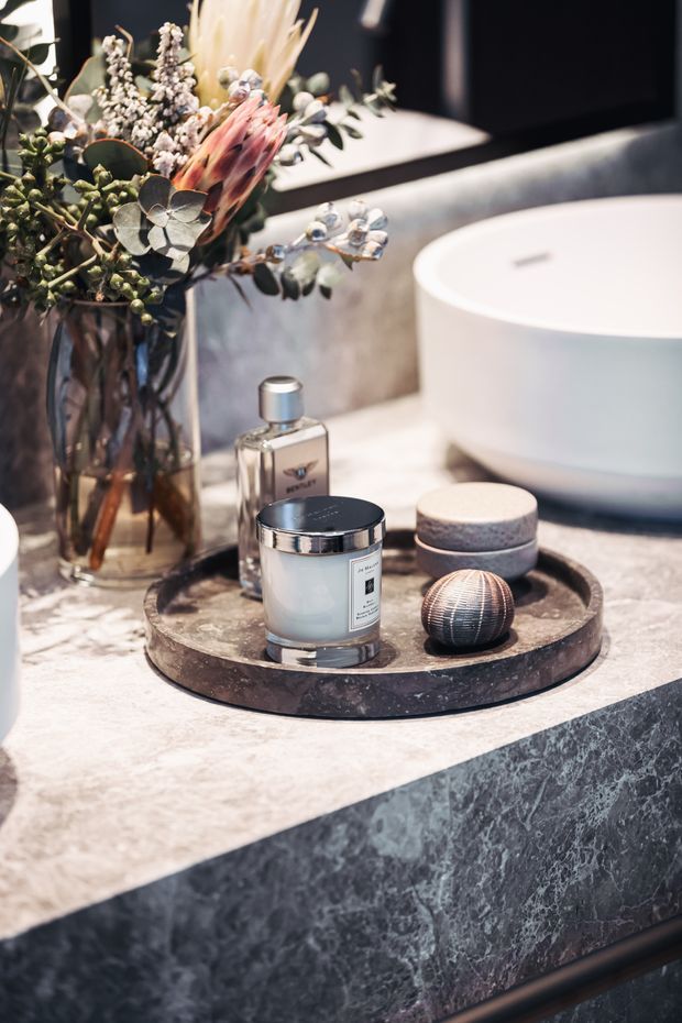 Close-up view of a refined bathroom vanity featuring a large white vessel basin, dark veined stone vanity surfaces, a black-framed mirror, and carefully arranged grooming and decorative accessories on a round tray. A floral arrangement adds an organic accent.