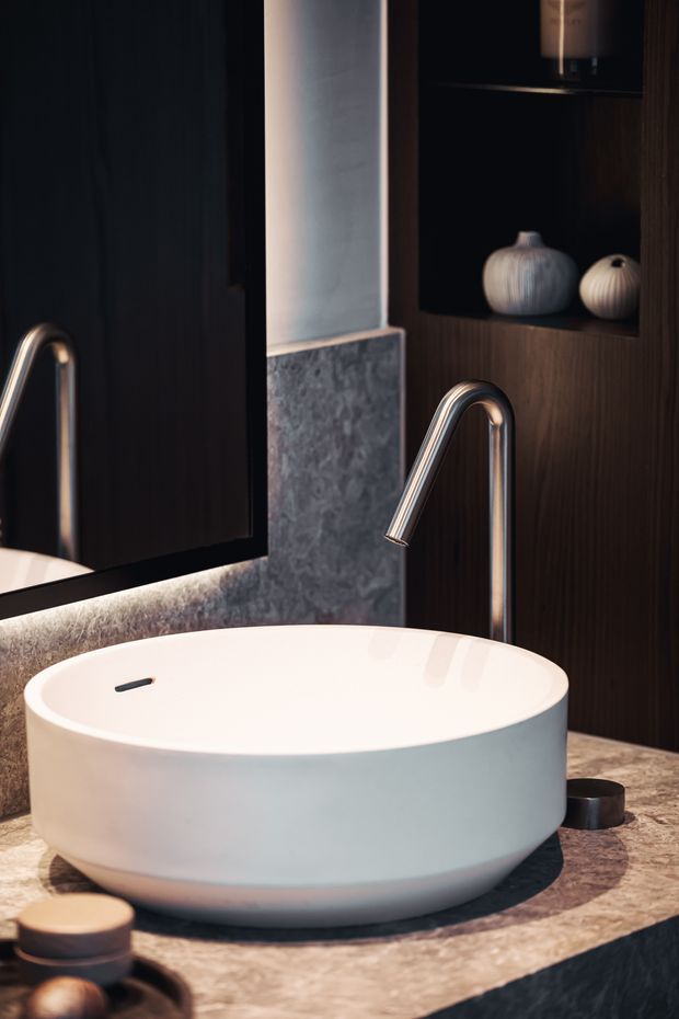 Close-up of a refined bathroom vanity featuring a freestanding round white vessel basin on a mottled natural-stone benchtop, with a tall brushed metal gooseneck tap. Dark timber wall cabinetry with open display shelving, ceramic vessels, and a large mirror create a moody, luxurious setting.
