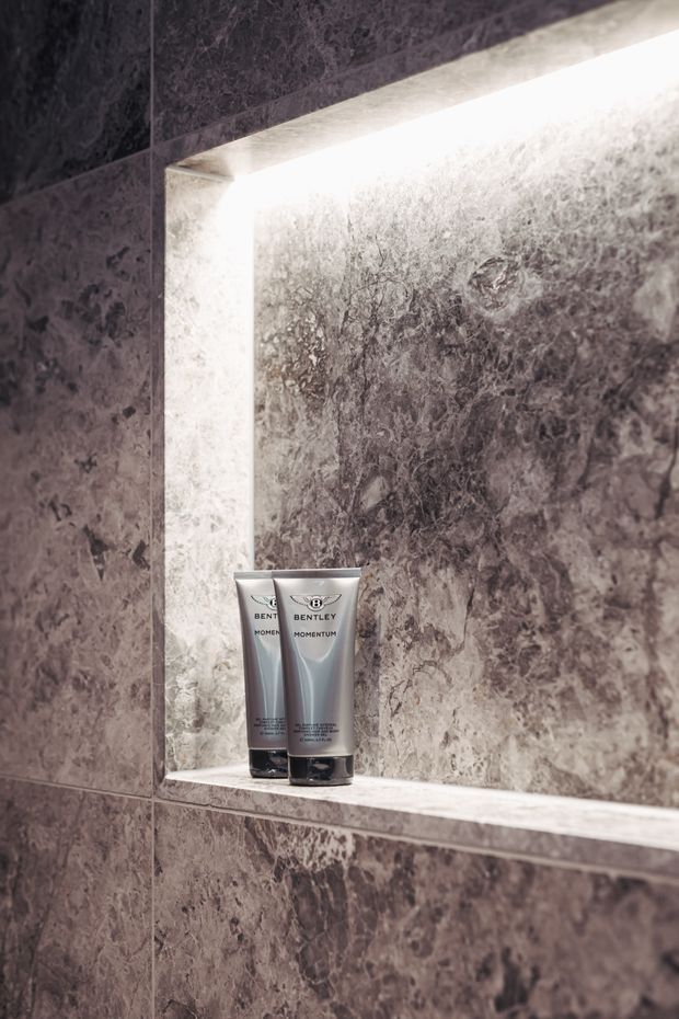 Close-up of a recessed, backlit wall niche lined with dark grey veined stone or marble-look slabs. Two toiletry tubes sit on the stone shelf, emphasizing the refined bathroom fit-out and integrated lighting.