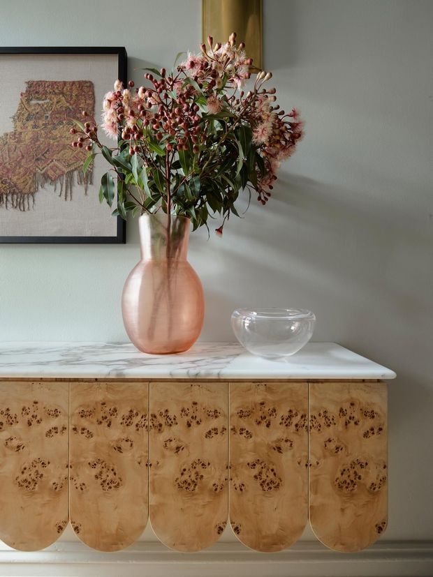 A styled interior vignette showing a warm timber sideboard with rounded cabinet doors and highly figured wood grain, topped by a white-grey marble surface. A blush glass vase filled with pink botanical branches and a clear glass bowl sit against a pale grey wall, with framed textile artwork partially visible behind.
