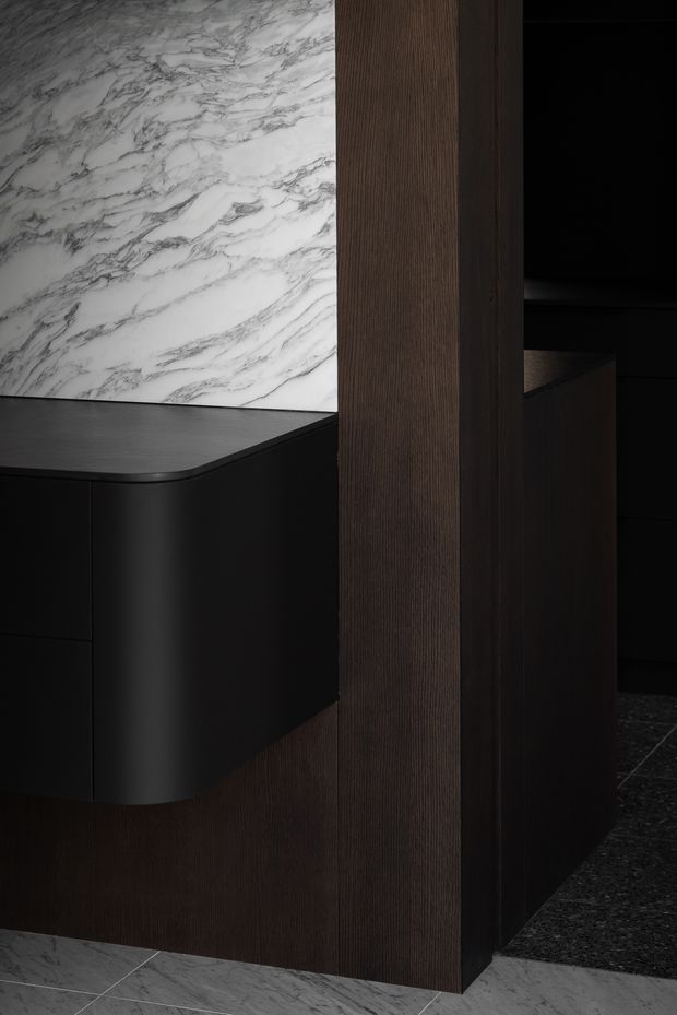 A dark, refined interior detail featuring a curved black built-in cabinet or vanity with a white marble-look veined stone splashback, framed by rich dark timber wall panelling. A second dark joinery element and charcoal flooring are visible in the background, creating a moody high-contrast composition.