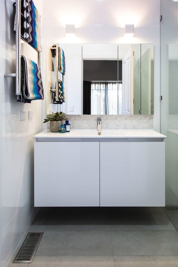 Bright contemporary bathroom featuring a wall-mounted white vanity with integrated basin, large mirrored cabinetry, a white herringbone tile splashback, grey tiled flooring, wall-mounted towels, and a glass-enclosed shower.