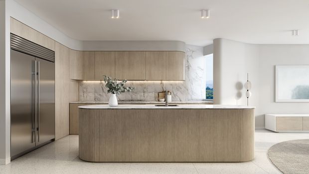 A bright, refined contemporary kitchen with a large curved island clad in vertically fluted light timber, matching curved wall cabinetry, a pale stone benchtop and backsplash, and integrated stainless-steel refrigeration. The open-plan space includes a glimpse of a living area, neutral furnishings, concealed lighting, and a large artwork.