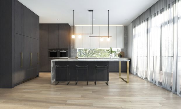 A spacious contemporary kitchen with a large island, dark full-height cabinetry, integrated appliances, pale timber flooring, and a light stone or concrete-look benchtop. Floor-to-ceiling sheer curtains line the right side, while a wide glazed opening above the rear bench brings in greenery and daylight. Linear pendant lighting and subtle brass-toned island supports add refined detail.