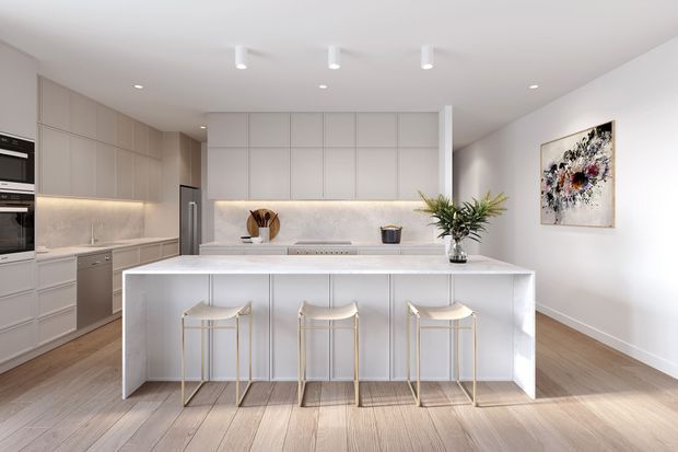 Bright contemporary kitchen with a large white island and three bar stools, full-height pale cabinetry, integrated appliances, a light stone-look splashback and benchtop, timber flooring, and recessed and track lighting. An adjacent open area with wall art is visible to the right.