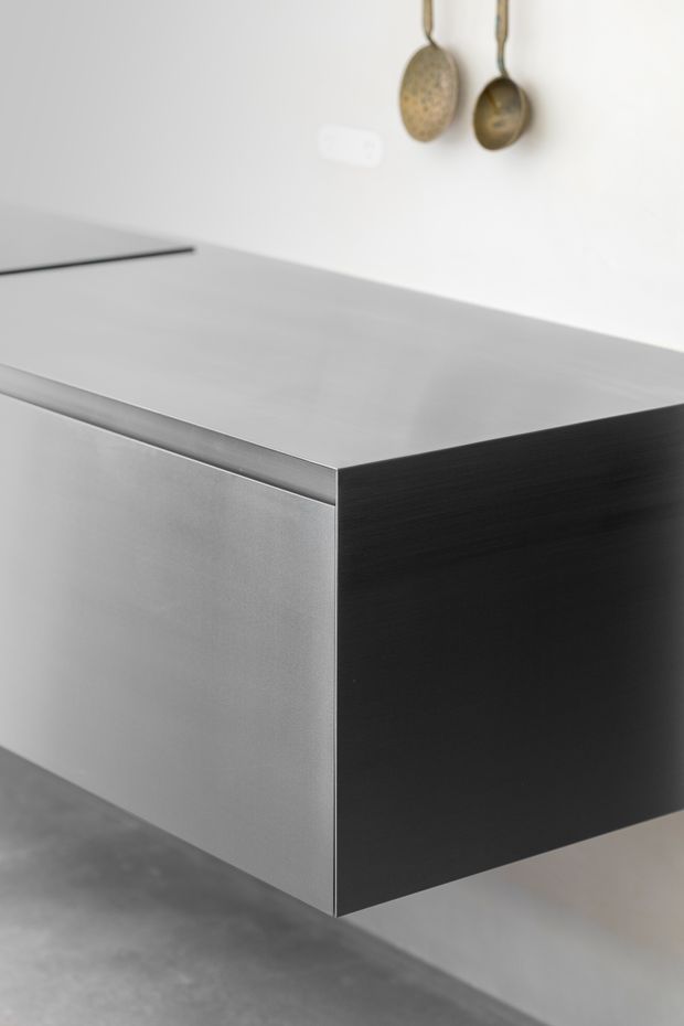 Close-up of a minimalist kitchen island or cabinet with a smooth brushed-metal benchtop and matching light grey metallic front, contrasted by a dark charcoal side panel. The island appears cantilevered or suspended above a grey floor. A white wall with two hanging metal utensils is visible in the background.