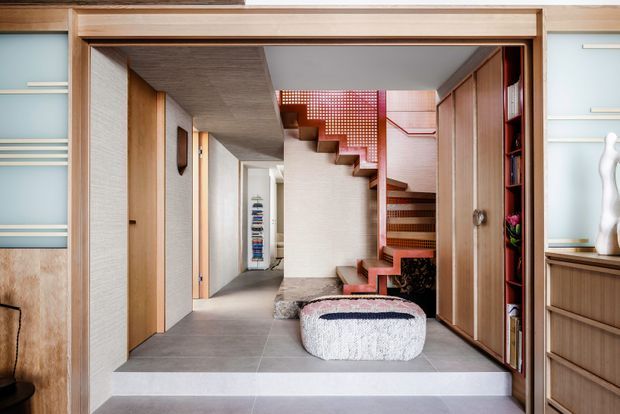 A refined residential hallway and stairway viewed through a wide timber-framed opening. The space features large grey floor tiles, pale textured wall finishes, extensive warm timber built-in cabinetry and doors, a sculptural upholstered ottoman, and a distinctive timber staircase with a perforated red-toned balustrade. Shelving and display joinery line the sides, creating a highly detailed contemporary interior.