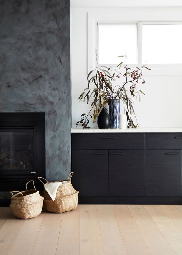 A refined contemporary interior featuring a black built-in fireplace beside a dark timber or stained-wood cabinetry wall. The cabinetry sits beneath a white-framed horizontal window and a pale benchtop or sill displaying decorative vases and branches. The left wall has a mottled dark grey feature finish, while wide pale timber floorboards and woven baskets add warmth and texture.