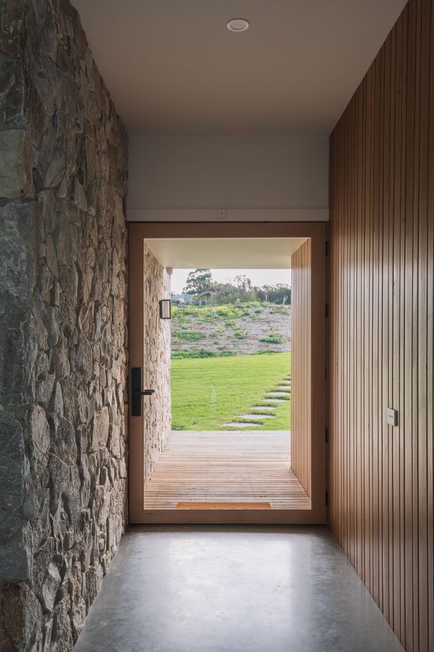 A narrow, contemporary entry hall leading through an open timber-framed doorway to a covered timber deck and landscaped garden. One wall is constructed from exposed irregular natural stone, while the opposite wall features vertical timber battens. The floor is smooth concrete, with a simple recessed ceiling light and dark door hardware. The view beyond shows lawn, stepping stones, and a lightly planted outdoor area.
