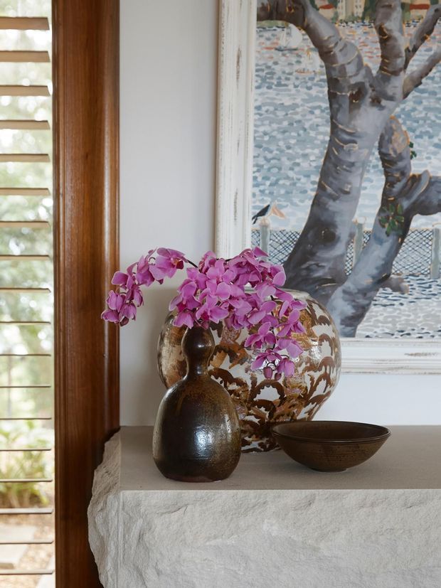 Close-up interior vignette of a textured white stone or plaster mantel beside a timber-framed window with horizontal shutters. A large patterned ceramic vessel filled with purple orchids, a dark rounded ceramic vase, and a shallow bowl sit in front of a framed artwork depicting a tree. The composition has warm natural materials and an eclectic, refined decorative character.
