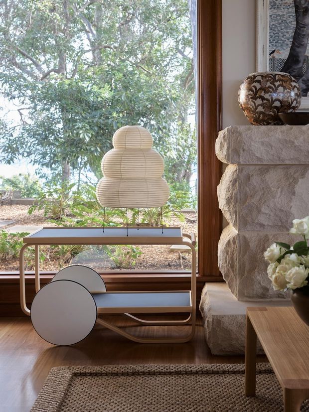 A warm, light-filled interior corner beside expansive timber-framed glazing, featuring a sculptural layered pendant lamp on a low console, a stone-clad fireplace or feature wall, a patterned ceramic vessel, flowers, and a timber coffee table over a woven rug. Mature greenery is visible outside through the window.
