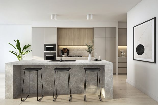 A bright contemporary kitchen centred on a large stone island with three black metal bar stools. The kitchen features pale full-height cabinetry, warm timber-look cabinetry around the cooktop, integrated black appliances, a light stone benchtop and matching waterfall island sides, pale timber flooring, white walls, and minimalist framed artwork. A small dining or living area is visible beyond the kitchen.
